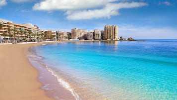 Une plage aux eaux turquoise et limpides, un rivage sablonneux et, en arrière-plan, une rangée d’immeubles à plusieurs étages.