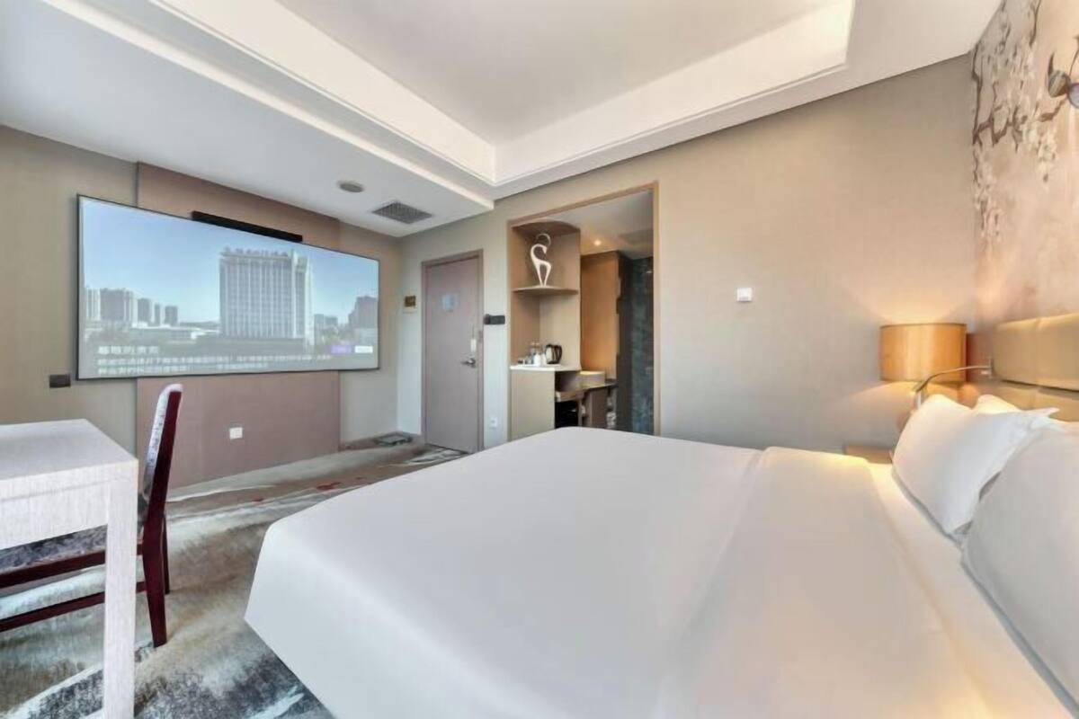 A hotel room with a large bed, a flat-screen TV mounted on the wall, a desk with a chair, and a small seating area with a lamp.