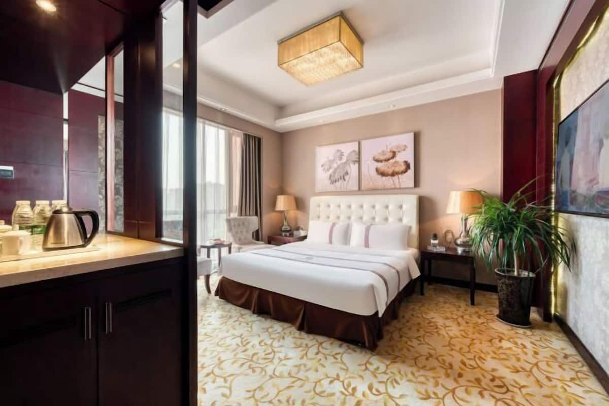 A hotel room with a large bed, a desk with a kettle, a chair, and a potted plant.