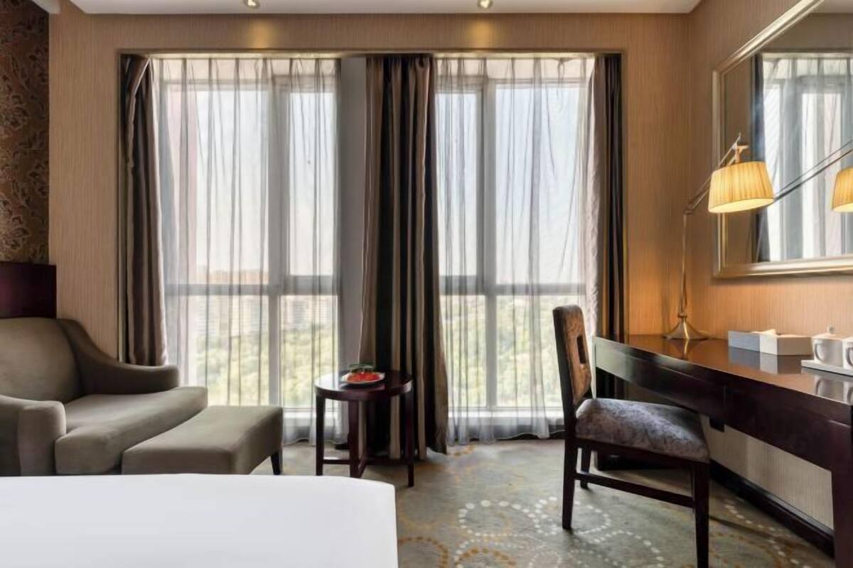 A hotel room with a bed, a desk, a chair, a lamp, and a view of the outdoors through large windows.