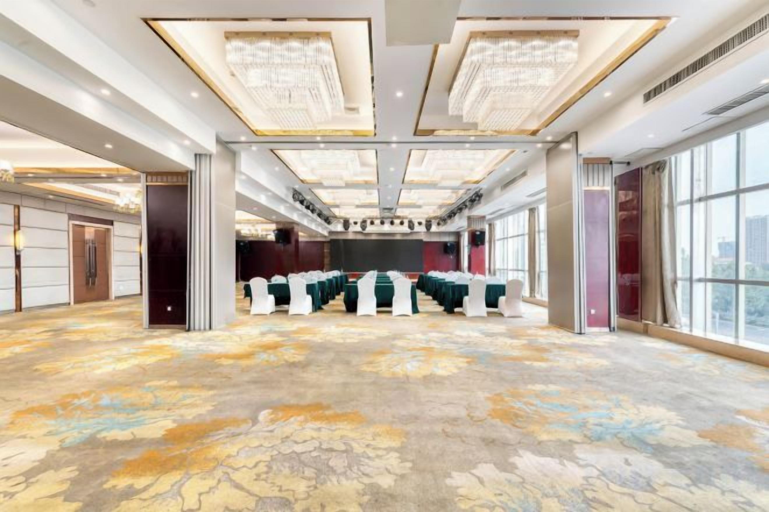 Un amplio salón de conferencias con alfombra estampada, techos altos con candelabros y ventanas grandes.
