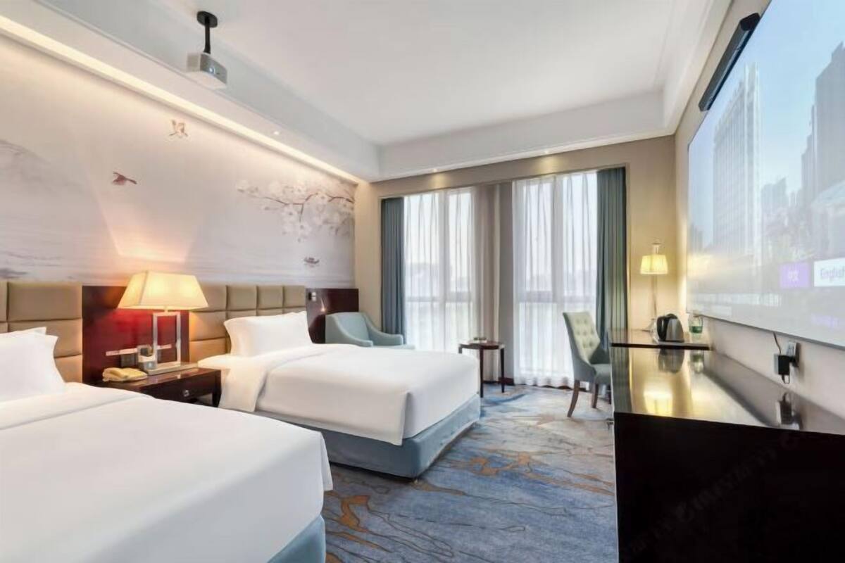 A hotel room with two beds, a desk, a chair, a TV, and a large window with curtains.