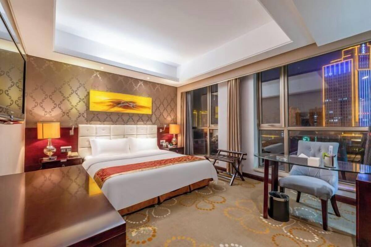 A modern hotel room with a large bed, a desk, a chair, and a view of the cityscape through the window.