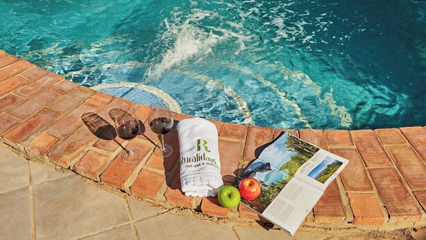 Escena junto a la piscina con una toalla, dos copas de vino tinto, una manzana, una revista y gafas de sol.