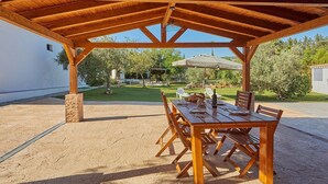 Un espacio de comedor al aire libre con techo, una mesa y sillas de madera, un pilar de piedra y vistas a un jardín y piscina al fondo.