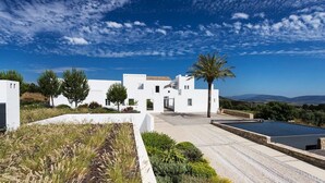 Une villa moderne aux murs blancs, dotée d’une piscine, entourée de verdure et surplombée d’un ciel bleu azur.