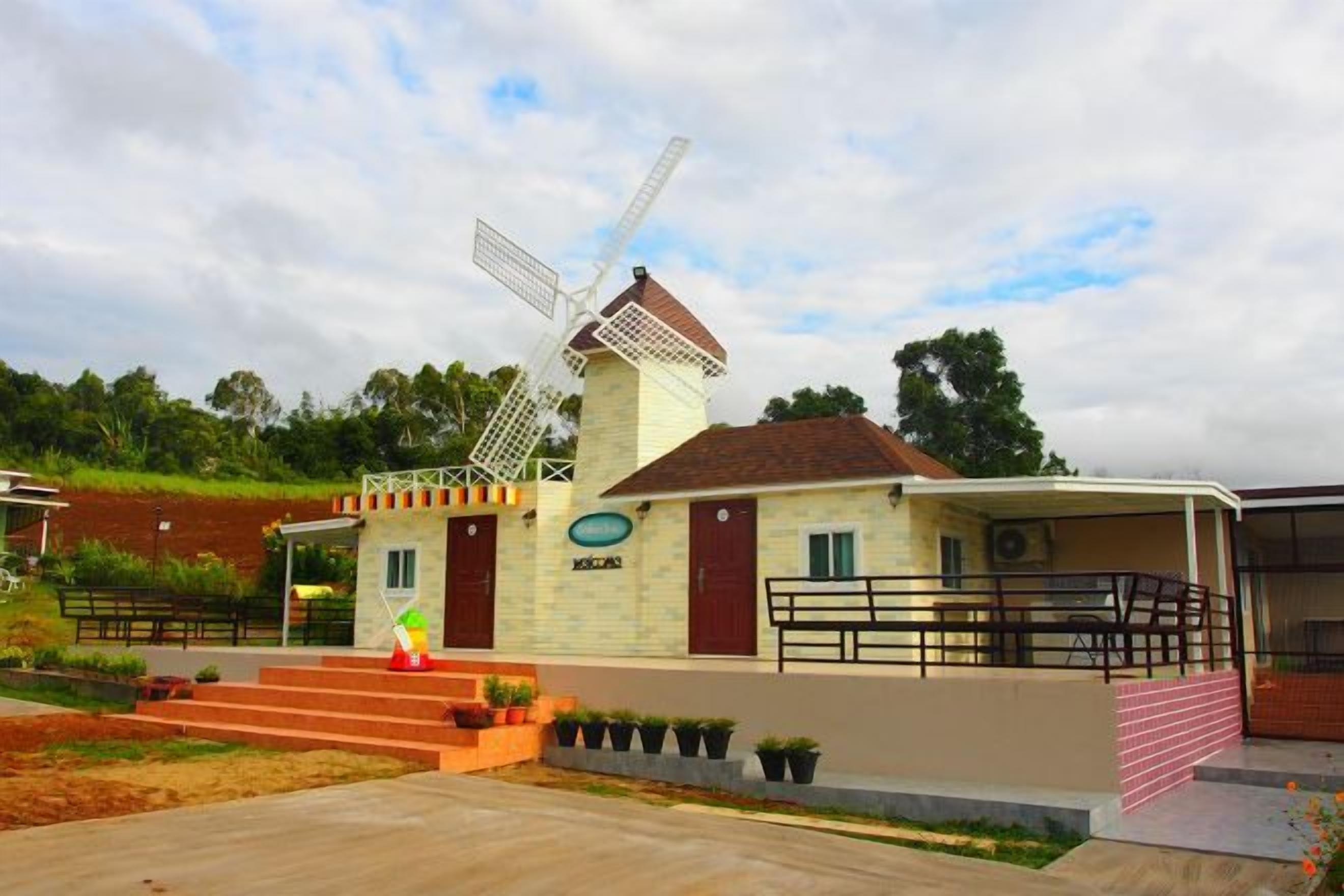 Um moinho de vento com base de tijolos e estrutura branca, um pequeno edifício com porta vermelha e uma área pavimentada com degraus que levam ao edifício.