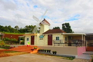 Un moulin à vent avec une base en briques et une structure blanche, un petit bâtiment doté d’une porte rouge, et une zone pavée avec des marches menant à l’entrée du bâtiment.