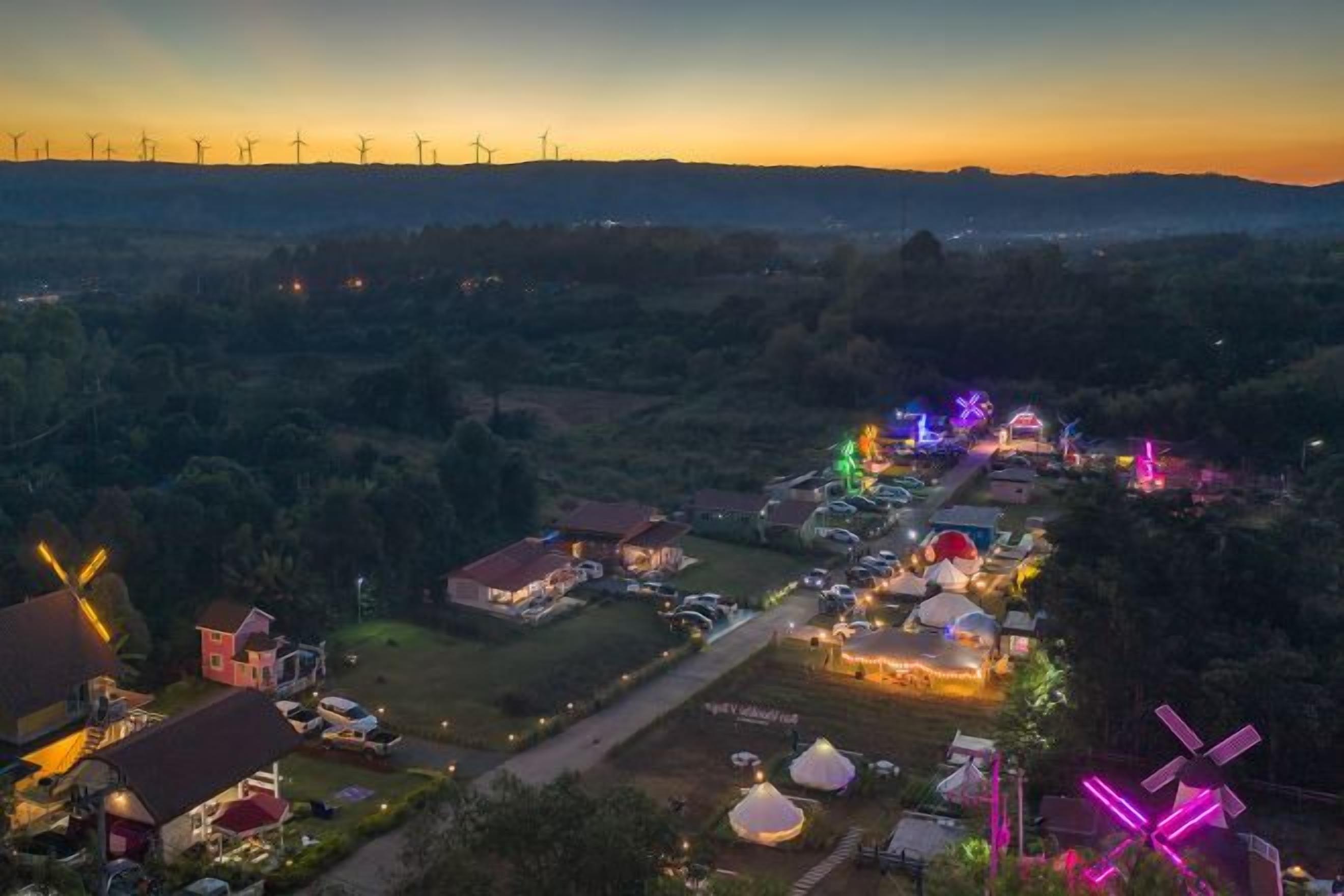 A rural area with wind turbines in the background, a lit-up village center, and a mix of residential and commercial buildings.