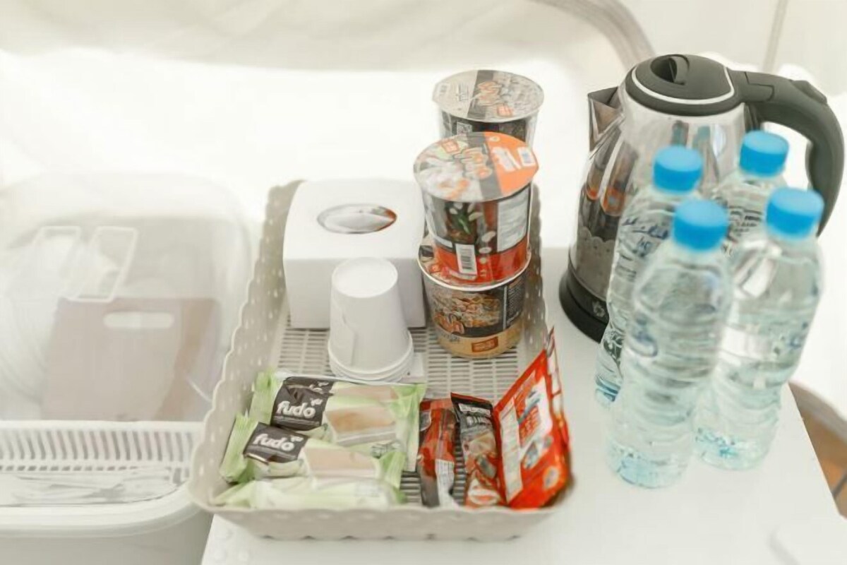 Een kitchenette met een waterkoker, waterflessen, een koffiezetapparaat en verpakte etenswaren.