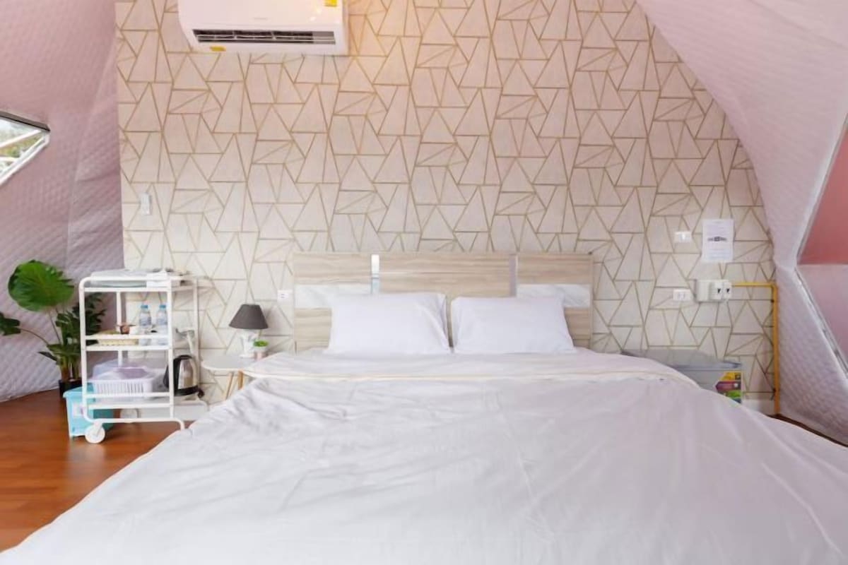 A bedroom with a geometric-patterned wall, a bed with white bedding, a bedside table with a lamp, and a small shelf with items.
