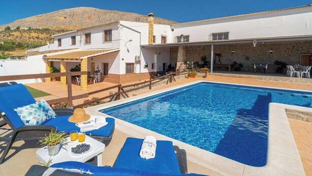 A poolside area with lounge chairs, a table with a drink and snacks, and a view of a building with a stone facade. - La Antigua Fábrica Teba by Ruralidays (Málaga)