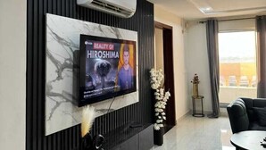 A modern living room with a mounted TV displaying 'Reality of Hiroshima', a black sofa, a dining table, and a decorative plant. - frnish flat  (Islamabad)