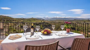 A table set for two with a bottle of wine, a fruit bowl, and a book on a balcony overlooking a mountainous landscape.