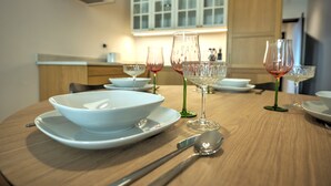 Una mesa de comedor dispuesta con platos, cuencos y copas de vino blancos, con una mesa de madera y una cocina al fondo.