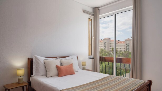 Un lit bien fait, recouvert d’une couvre-lit à rayures, avec deux lampes de chevet et une vue sur les bâtiments par une grande fenêtre.