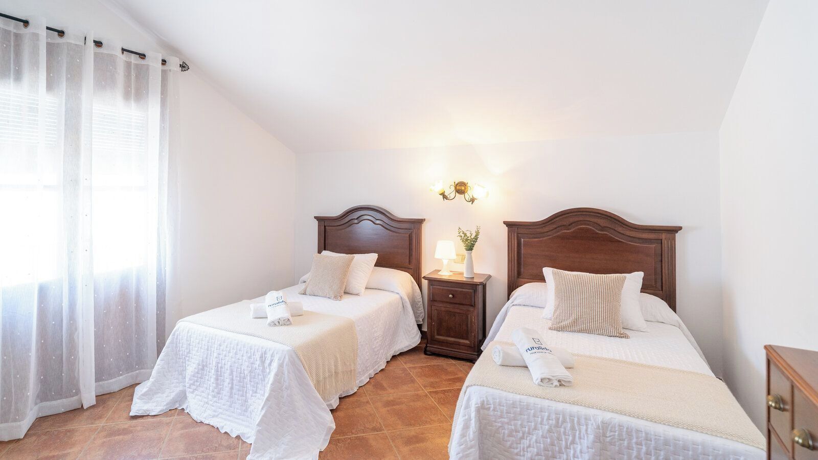Quarto com duas camas de solteiro, uma mesa de cabeceira com abajur e uma janela com cortinas leves.