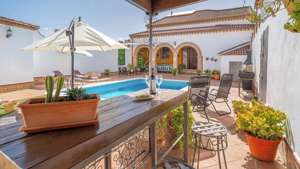 Uma área à beira da piscina com mesa de madeira, cadeiras e plantas em vasos.