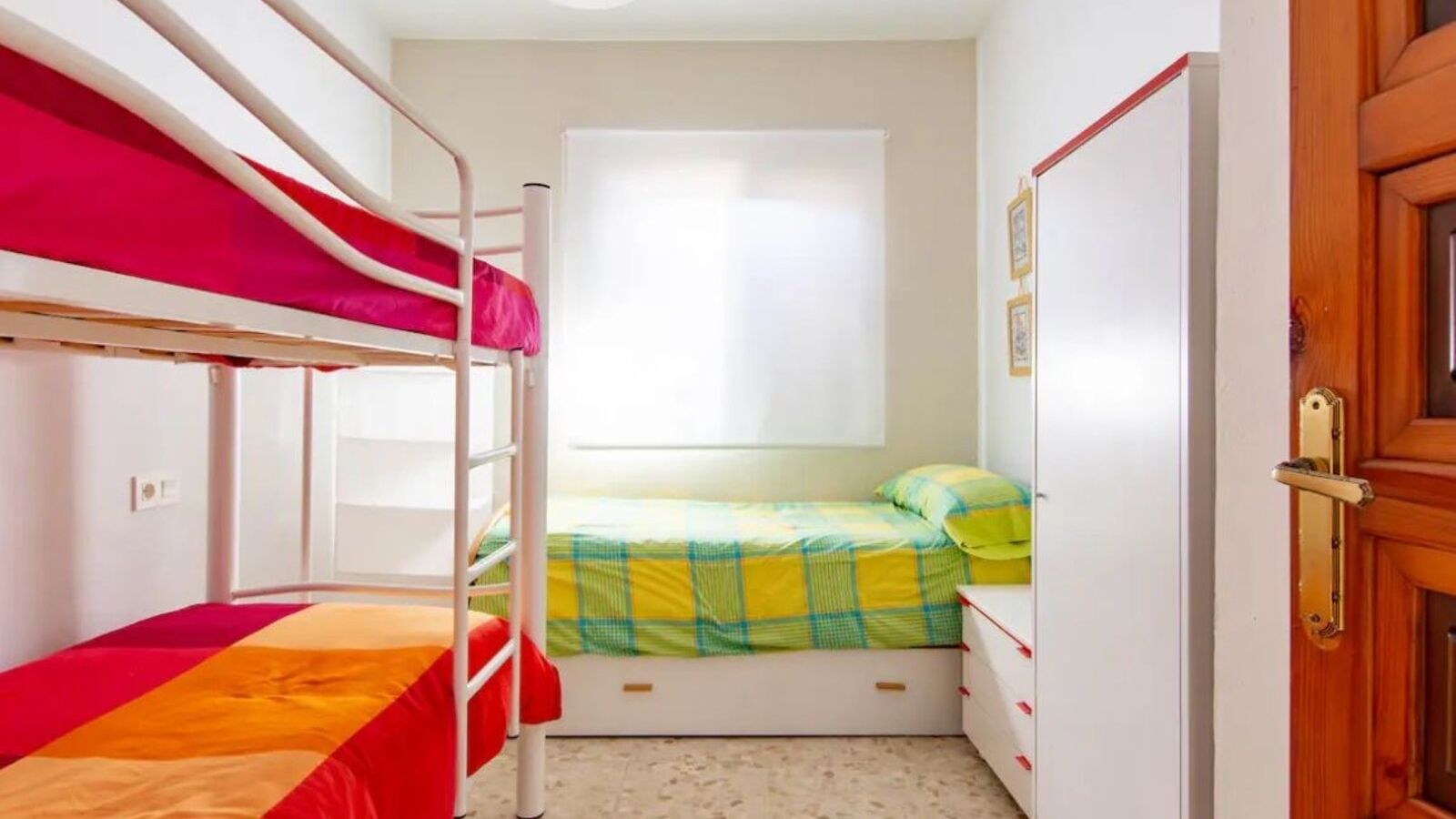 Een slaapzaal met stapelbedden, een eenpersoonsbed, een raam, een deur en een kleine dressoir.