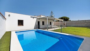 Una piscina rectangular con una terraza blanca, rodeada por un muro blanco y una zona de césped.