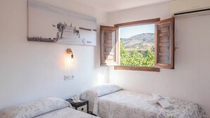 Un dormitorio con dos camas, una mesita de noche, una lámpara de pared y una ventana con vistas a zonas verdes y colinas.