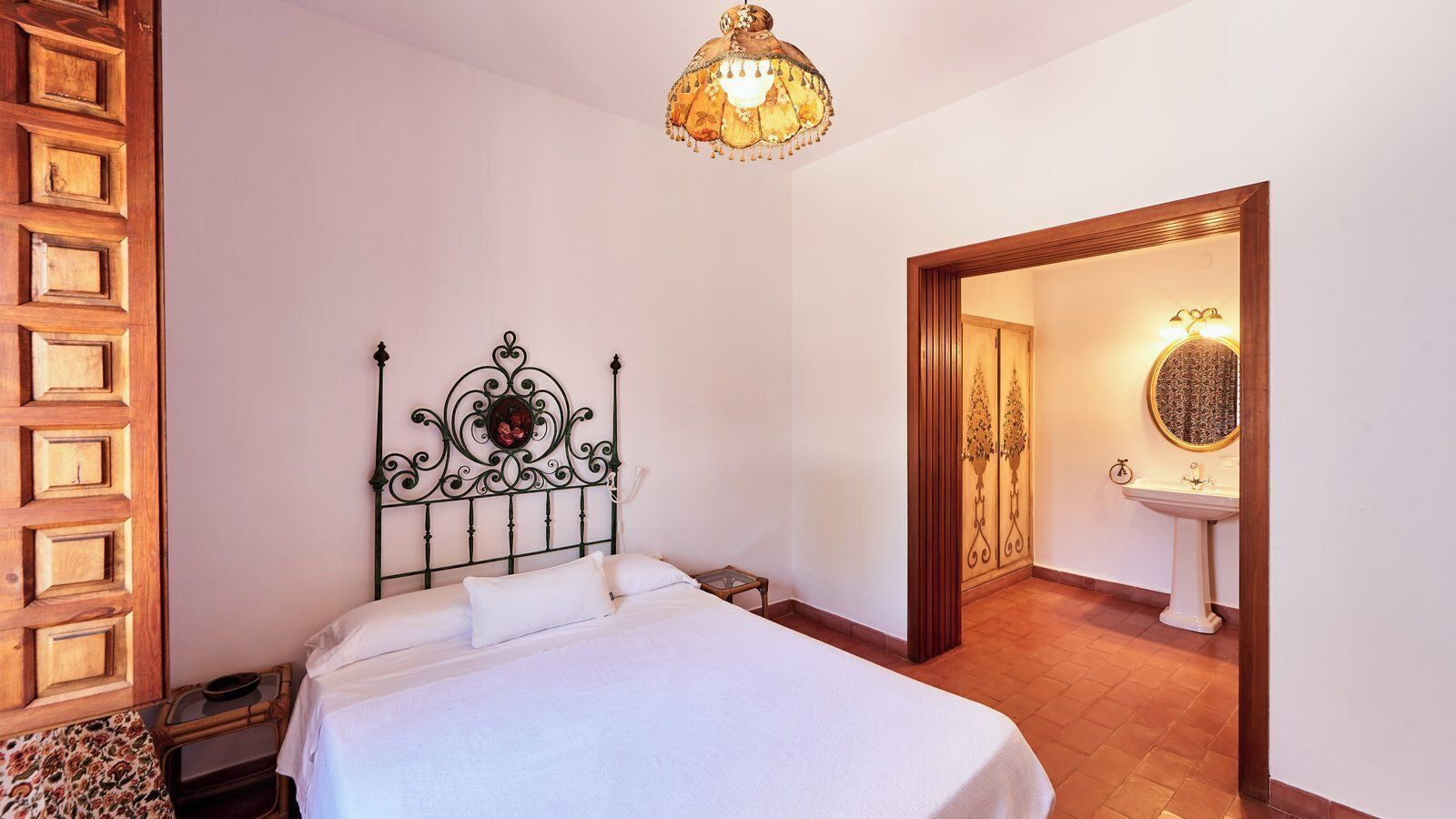 Un dormitorio con cama, cabecera decorativa, mesita de noche y teléfono en la pared, y una puerta que da a un baño con lavamanos y espejo.