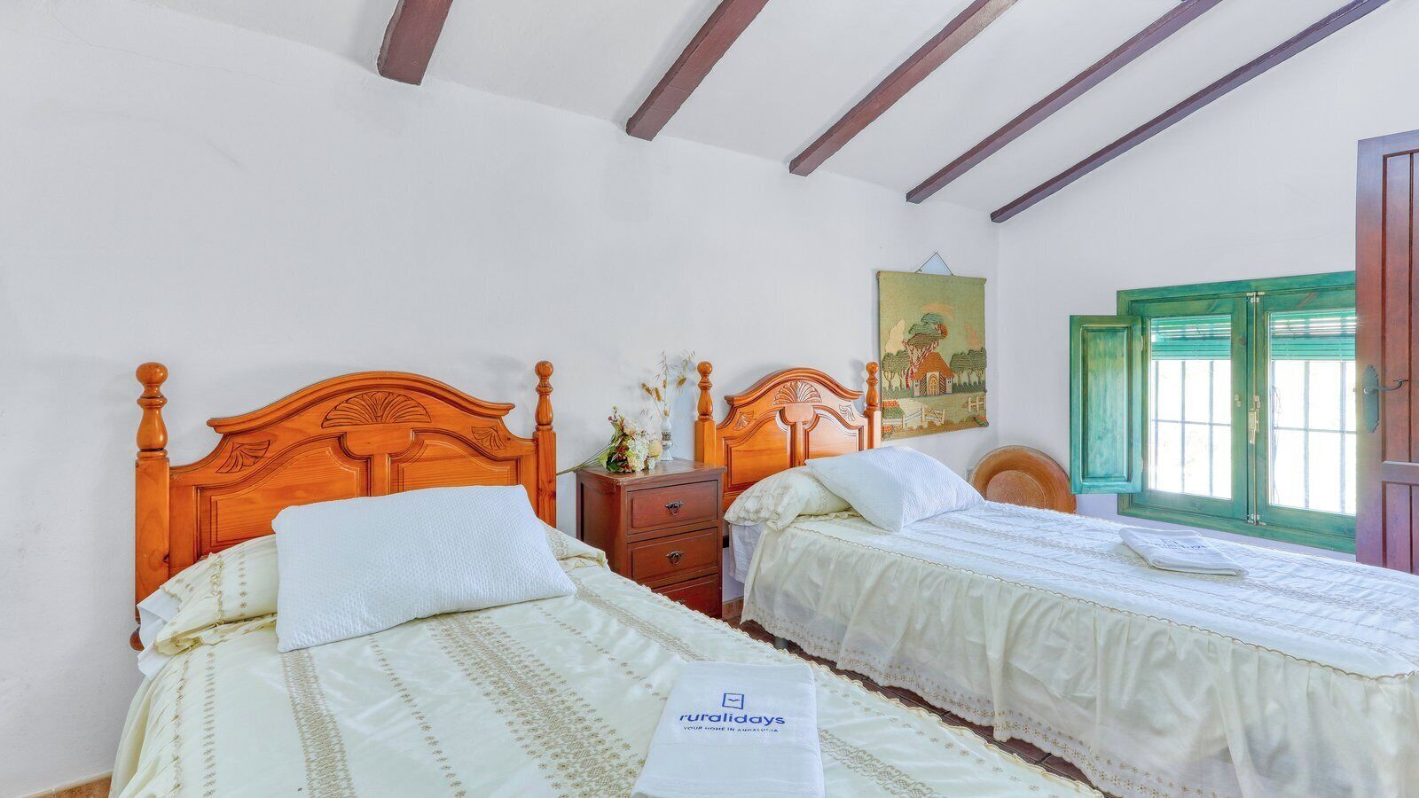 A bedroom with two wooden beds, a nightstand, a painting, and a window with green shutters.