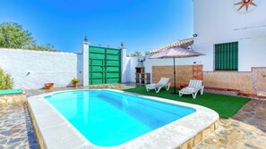 Una piscina rectangular con una reja verde, rodeada por un muro blanco y un patio con baldosas que incluye dos sillas reclinables blancas y un paraguas morado.