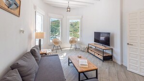 En moderne stue med sofa, sofabord i træ, fletstole, et fjernsyn og store vinduer med udsigt til grønne områder.
