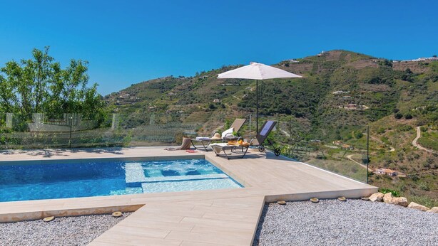 En swimmingpool med liggestole og parasol, omgivet af en stenstrand med udsigt til et bølgende landskab.