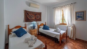 A room with two single beds, a nightstand with a lamp, a tapestry on the wall, and a window with curtains.
