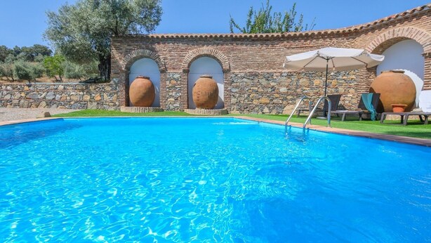 A swimming pool with stone walls and arched openings, surrounded by a grassy area and trees. - Cortijo San Rafael Constantina by Ruralidays (Seville)