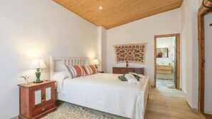 Un dormitorio con techo de madera, una cama con ropa de cama blanca, una mesita de noche con lámpara, un armario de madera y un tapiz en la pared.