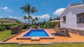 Una piscina rectangular con borde de piedra, rodeada por una terraza con sillas de descanso y una sombrilla.