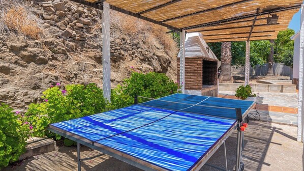 Una mesa de ping pong al aire libre ubicada bajo una pérgola de madera, con un muro de piedra y follaje en el fondo.