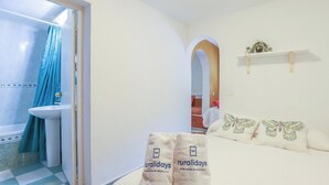 Un baño con lavamanos y ducha, una cama con almohadas de mariposa, y una repisa con una planta.