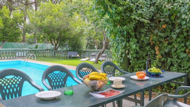 Zona de piscina con mesa lista para comer, rodeada de sillas y vista a un jardín con árboles y una valla.