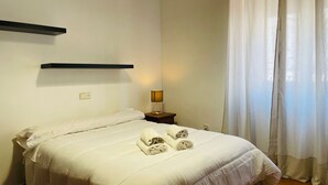 Un letto rifatto con lenzuola bianche e asciugamani arrotolati, una lampada da comodino e una finestra con tende leggere.