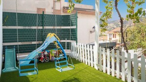 Un parque infantil con un tobogán y columpios, rodeado por una valla de piquetes blancos y una pared de escalada verde.