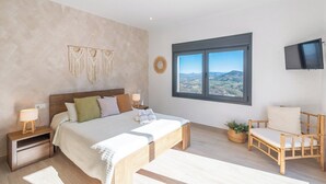 A bedroom with a large bed, a bedside table with a lamp, a wooden chair, a window with a view, and a TV on the wall.