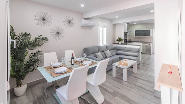 Una sala de estar moderna con comedor, sofá gris, sillas blancas y una pequeña cocina al fondo.