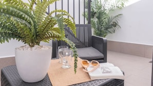 Un balcón con una planta en maceta, dos sillas, una mesa con vasos y un libro, y una alfombra pequeña.