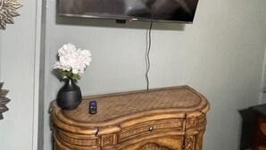 A flat-screen TV mounted on a wall above a wooden cabinet with a vase of white flowers. - Charming 3-bedroom house with AC and WiFi in peaceful Rockingham (Rockingham)