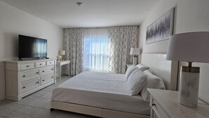 A modern hotel room with a large bed, a flat-screen TV, a dresser, and a bedside table with a lamp. - New Construction with Heated Swim Spa 5,5 mi to the beach with fenced back yard (Port Charlotte)
