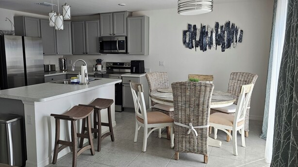 A modern kitchen with grey cabinets, stainless steel appliances, a central island with stools, and wicker chairs around a dining table. - New Construction with Heated Swim Spa 5,5 mi to the beach with fenced back yard (Port Charlotte)