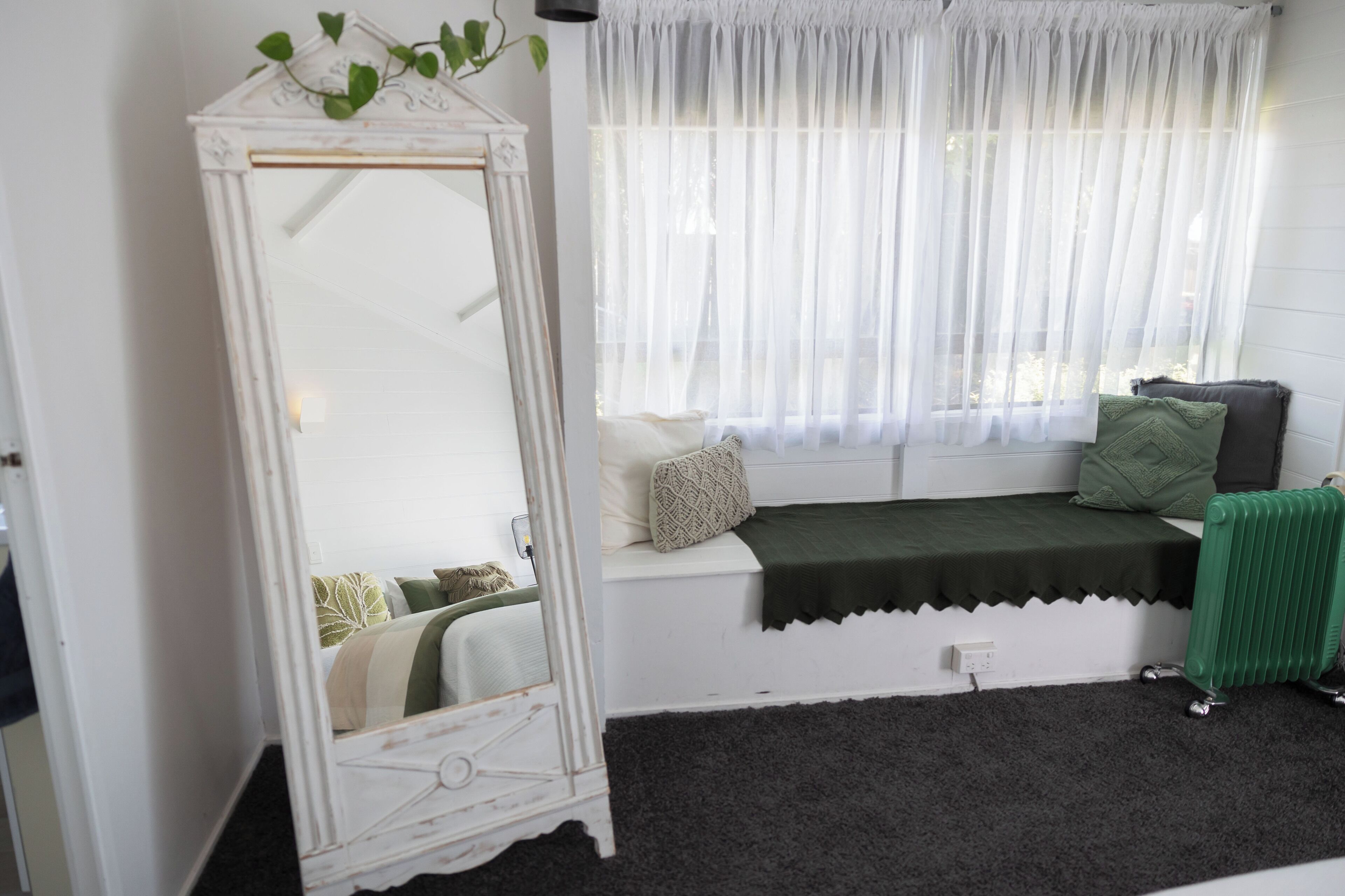 Una habitació amb un tocador blanc, una finestra amb cortines translúcides i un radiador verd, i una banca amb coixins.
