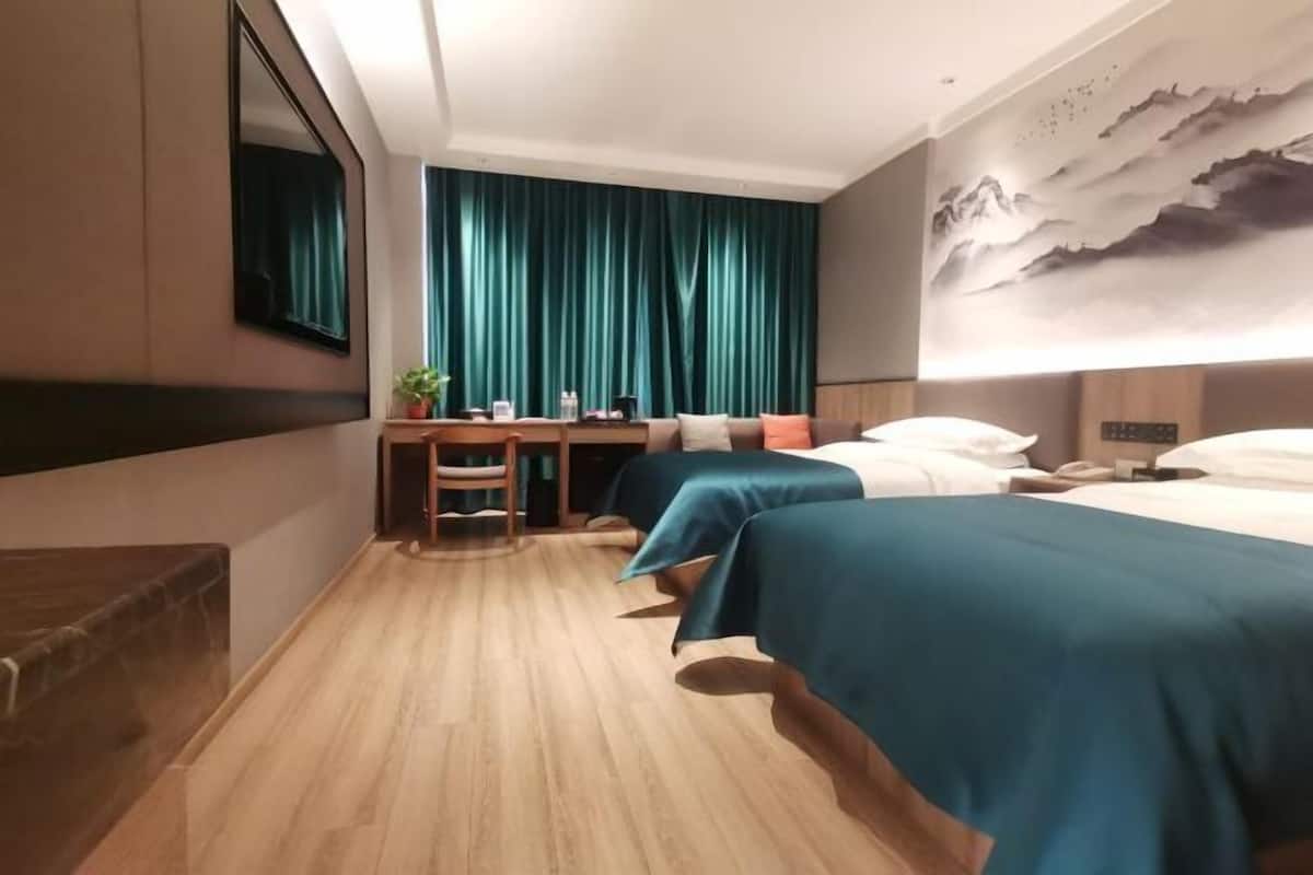 A hotel room with a wooden floor, a desk with a chair, two beds with teal bedspreads, a large painting on the wall, and a window with teal curtains.