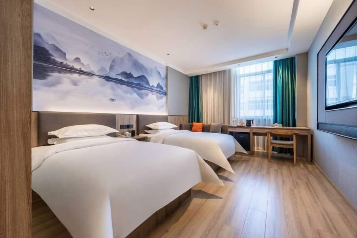 A hotel room with two beds, a desk, a chair, a large window, and a painting on the wall.