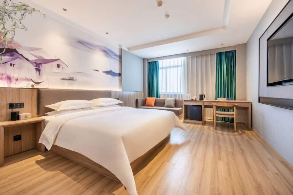 A modern hotel room with a large bed, a desk, and a chair, a TV mounted on the wall, and a painting on the wall.