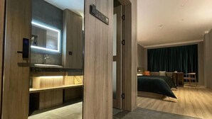 Una habitación de hotel moderna con detalles de madera, un lavamanos integrado, una cama con colcha verde oscuro y un pequeño rincón de descanso con una silla y una mesa.